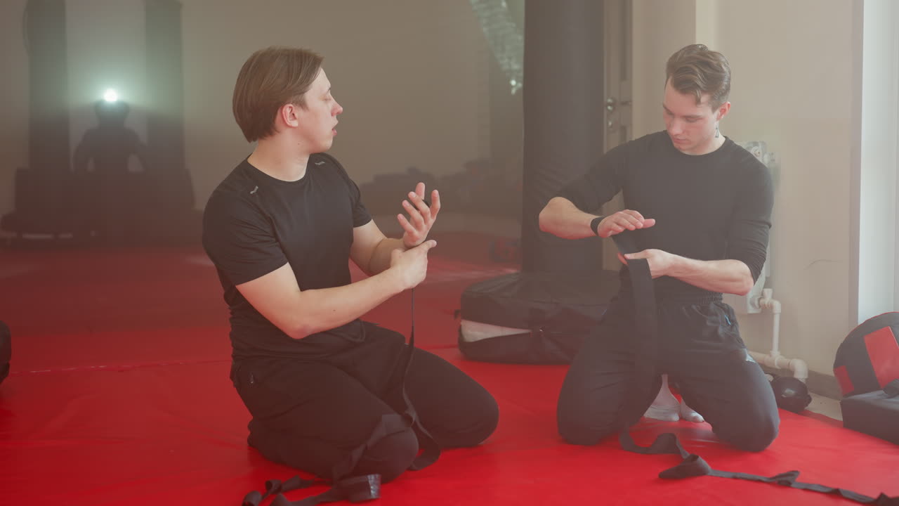 Two athletes kneel on red mat preparing hands with black wraps before workout, focusing on technique and discipline in combat sports training environment, showing teamwork, strength, for martial arts practice