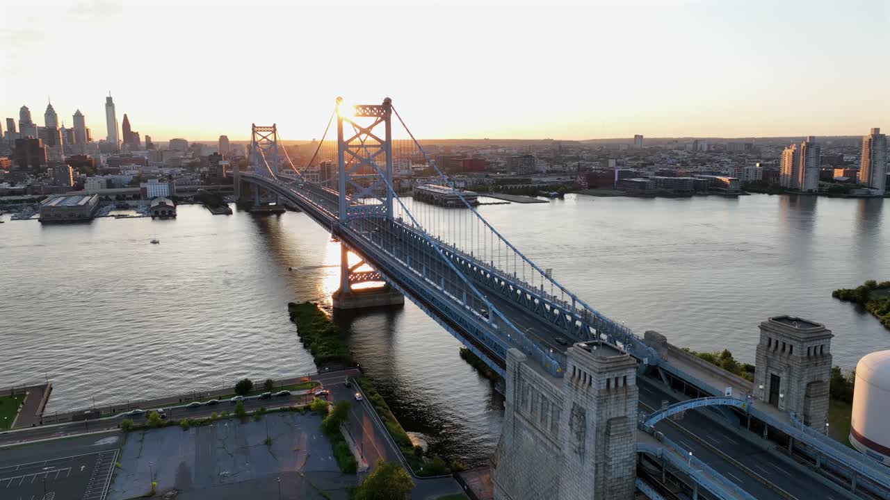 Aerial orbiting shot of iconic Benjamin Franklin bridge between Camden and Philadelphia. Sunset time behind skyline and skyscrapers of philly. Wide shot. Delaware River between two states