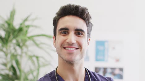 Video-portrait-of-happy-biracial-male-doctor-smiling-in-hospital-corridor,-with-copy-space