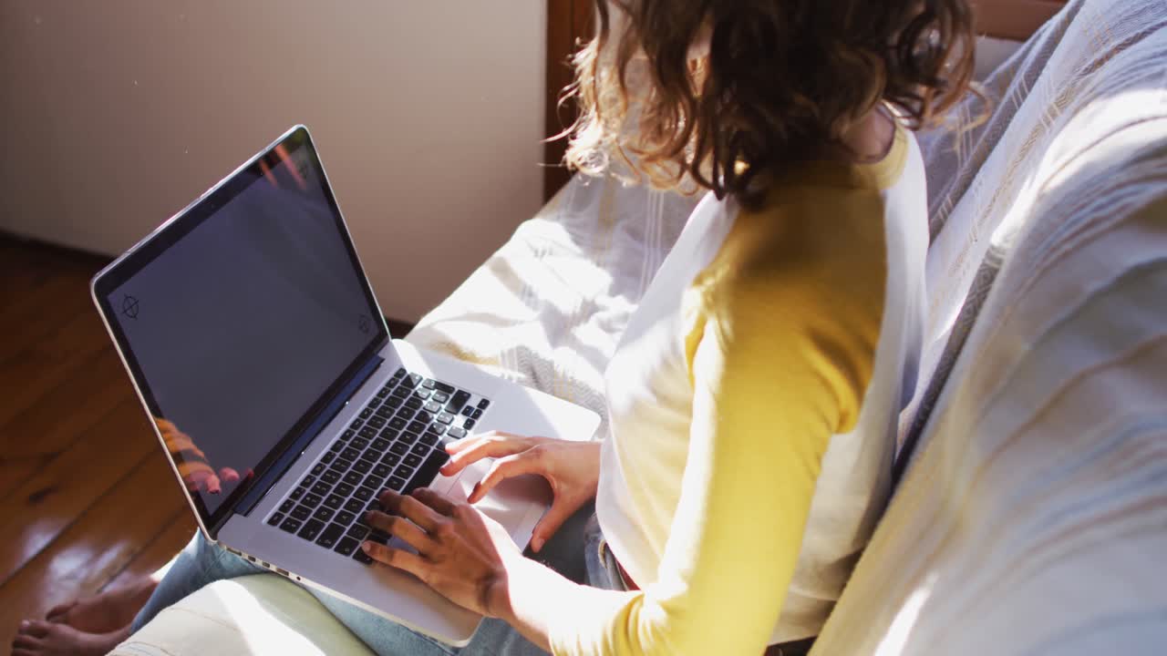 mujer de raza mixta sentada en el sofá usando una computadora portátil en la sala de estar soleada