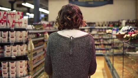 Rare-View-Of-A-Woman-With-Wavy-Hair-Is-Driving-Shopping-Trolley-Through-Food-Department-In-Supermarket-And-Looking-Around