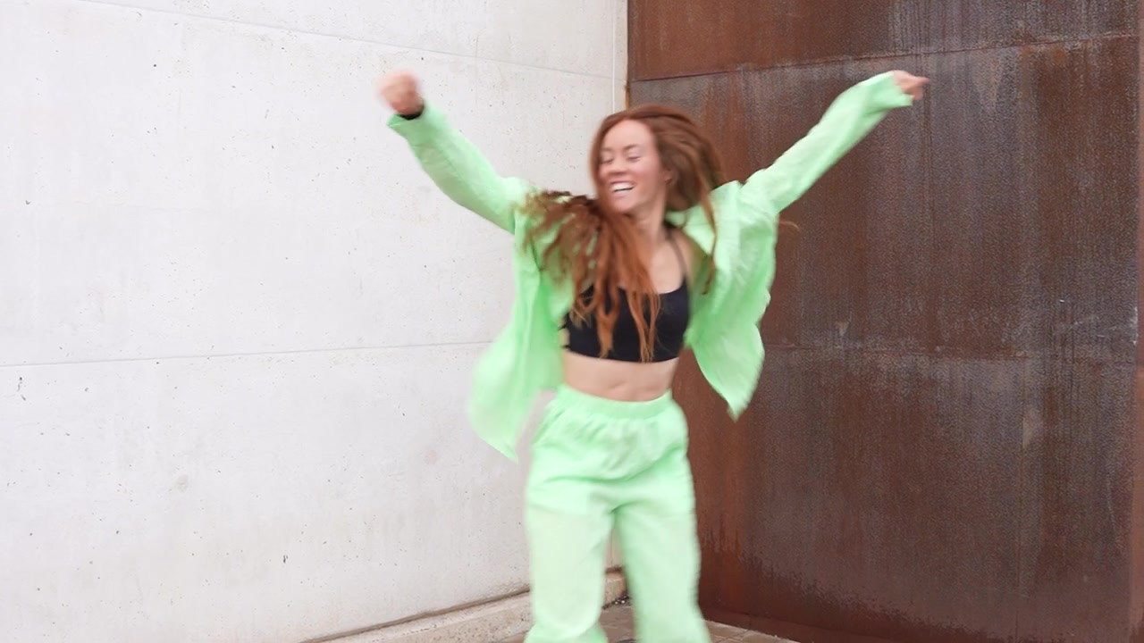 A woman is energetically dancing outdoors in a lime green suit with a modern urban backdrop