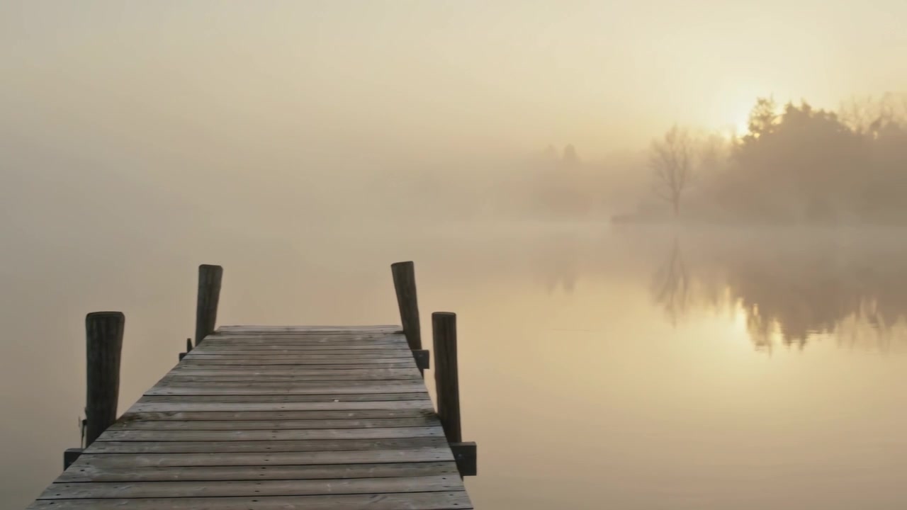 Jutting worn pier pointing into foggy lake as sun rising lifting mist, revealing tree reflection