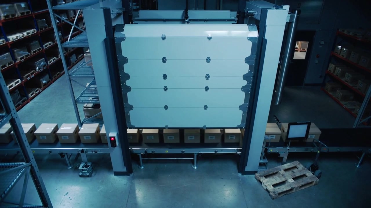 Moving conveyor carrying boxes under lowering panel in warehouse, pallet entering causing sealing
