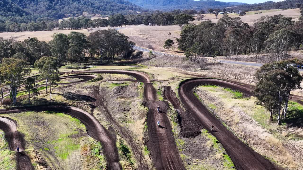 Dirt bikes race along a winding outdoor motocross track under bright daylight, aerial perspective