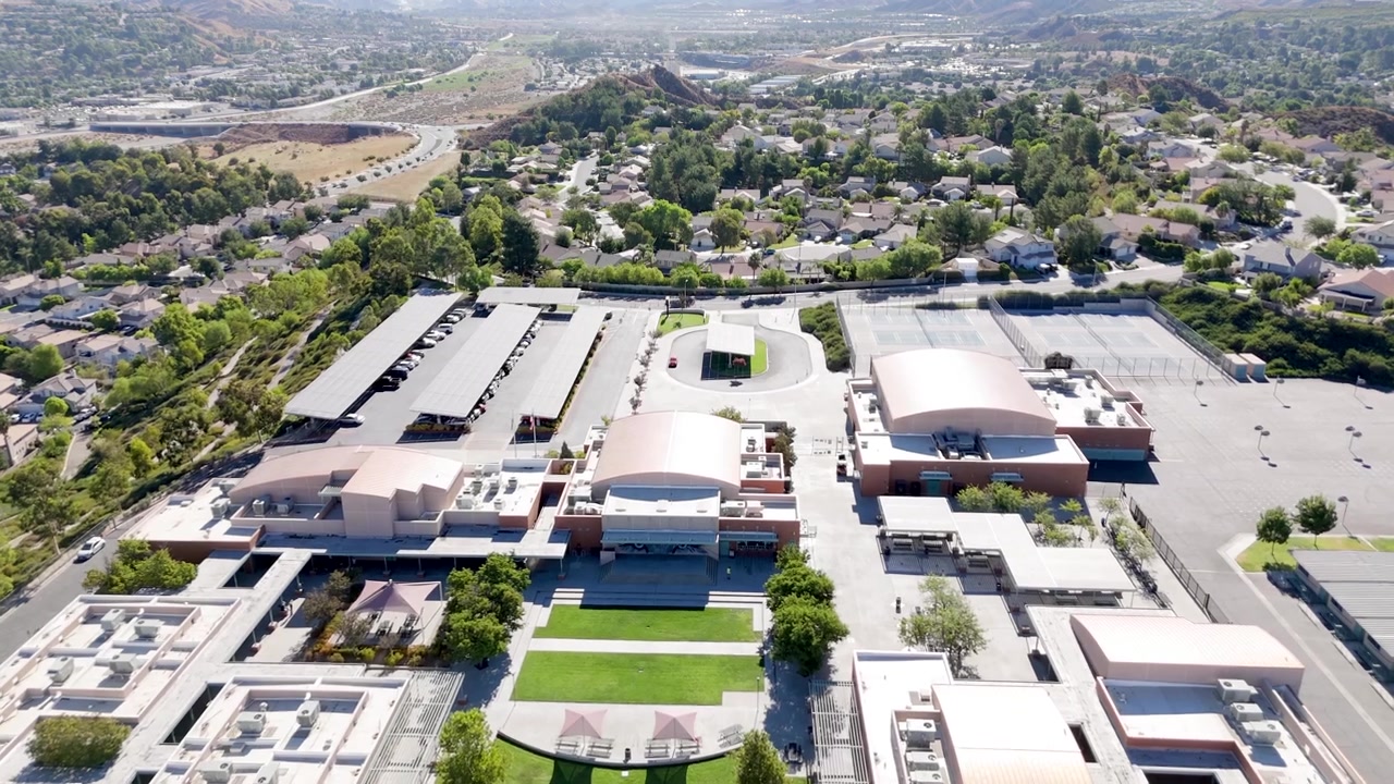 Sobrevoo de drone de um campus moderno de escola secundária em Santa Clarita, Califórnia, apresentando arquitetura contemporânea, estacionamento coberto, campos esportivos e espaços verdes vibrantes em plena luz do dia