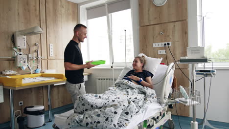 caring husband bring food to his wife in hospital. pregnant lady lying in the bed smiles to her man.