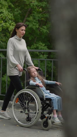 mom walks on pavement with little daughter sleeping in wheelchair. adult woman takes girl with muscular dystrophy out to city park on spring day