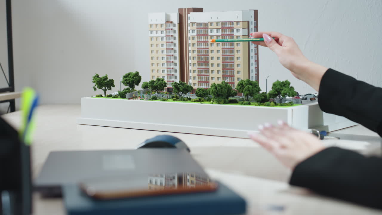 Business expert gesturing with pencil while explaining details of modern residential building model during office meeting, with green trees in model foreground