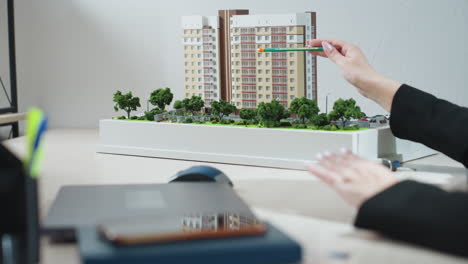business expert gesturing with pencil while explaining details of modern residential building model during office meeting, with green trees in model foreground