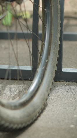 primer plano de una rueda de bicicleta estacionada con seguridad en un soporte de bicicleta de metal, enfatizando los radios y el marco, con detalles borrosos de las bicicletas circundantes y la vegetación en el fondo