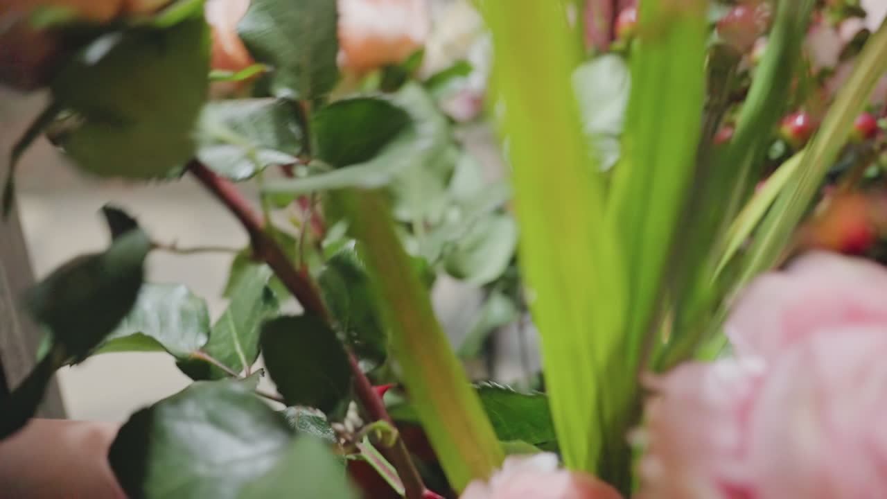Dettagli di una composizione floreale. Mani che sistemano steli in un vaso rosso con verde e rose visibili. Vista dettagliata della composizione floreale con mani che posizionano fiori in un vaso quadrato su un pavimento piastrellato riflettente.