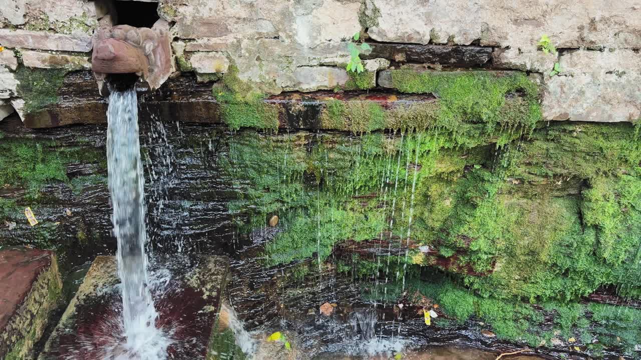 Close-up of water flowing from an intricately carved stone spout embedded in an old wall, channeling natural spring water with moss-covered textures and traditional craftsmanship