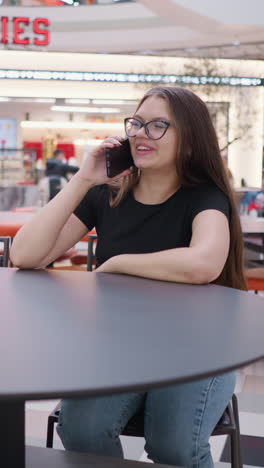 joven felizmente haciendo una llamada telefónica sentada en una mesa en un centro comercial ocupado, biggies señal visible por encima, con compradores y comensales alrededor