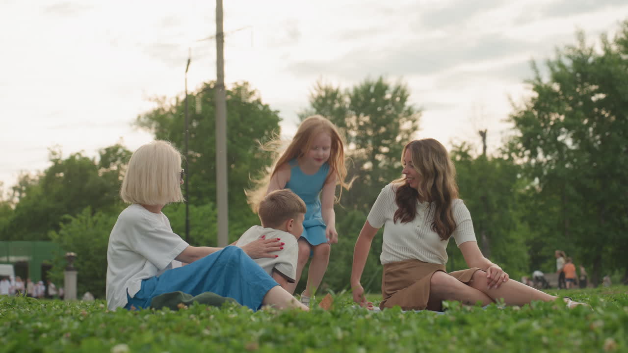 joyful moms sitting on mat in park calling running kids to join, playful energy and laughter, green grass background, casual summer gathering with children approaching for shared moment together