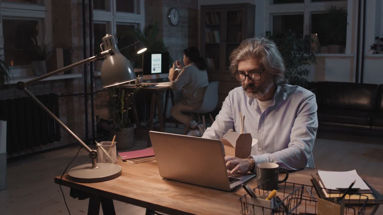 Grey Haired Man With Eyeglasses And Blue Shirt Sitting In Front Of Computer While Eating Food And Working, In The Background His Partner Works Sitting At A Desk
