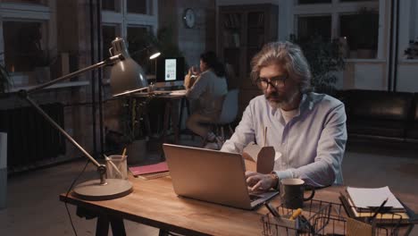 grey haired man with eyeglasses and blue shirt sitting in front of computer while eating food and working, in the background his partner works sitting at a desk