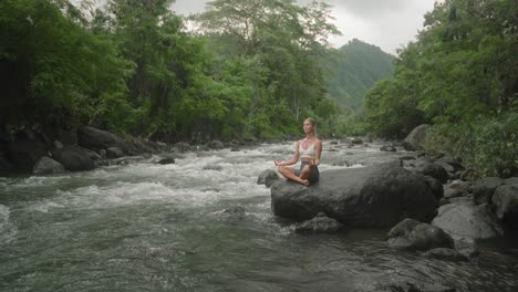 soul searching attractive female in lotus pose on rock along calming river