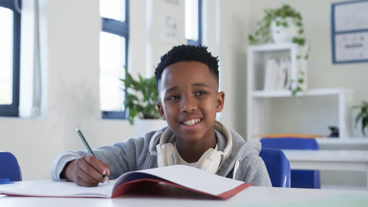 Adolescente africano seduto alla scrivania che scrive con una matita su un quaderno rosso, studia con cuffie e felpa grigia con cappuccio