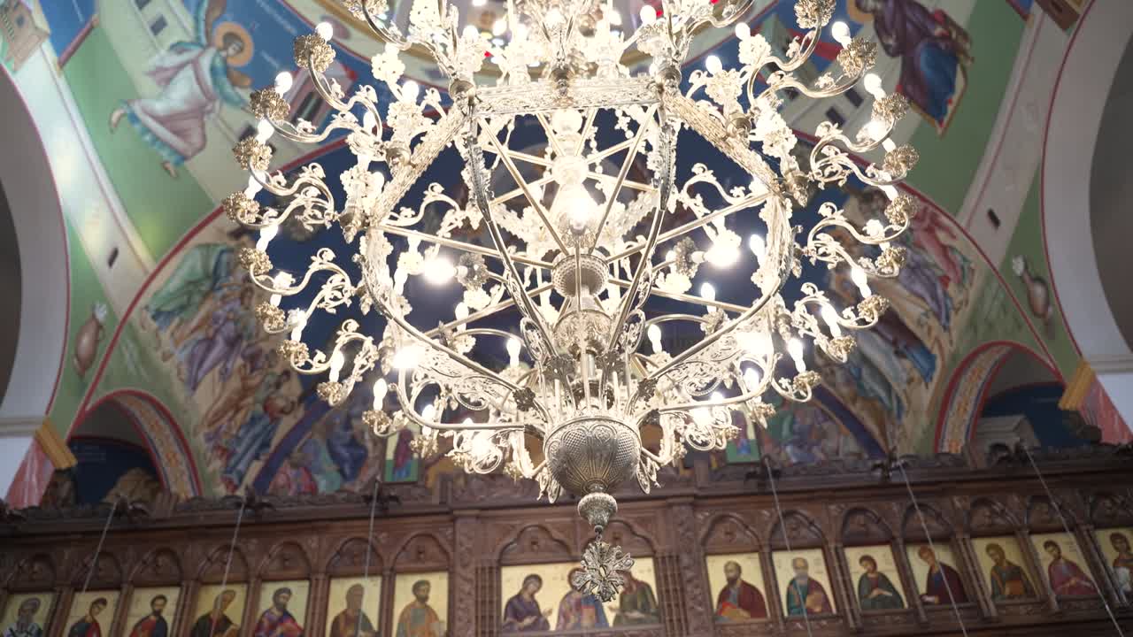 The camera shows a chandelier inside a church. Colorful paintings and icons decorate the walls. Light shines from the chandelier. People may visit this place for worship and events
