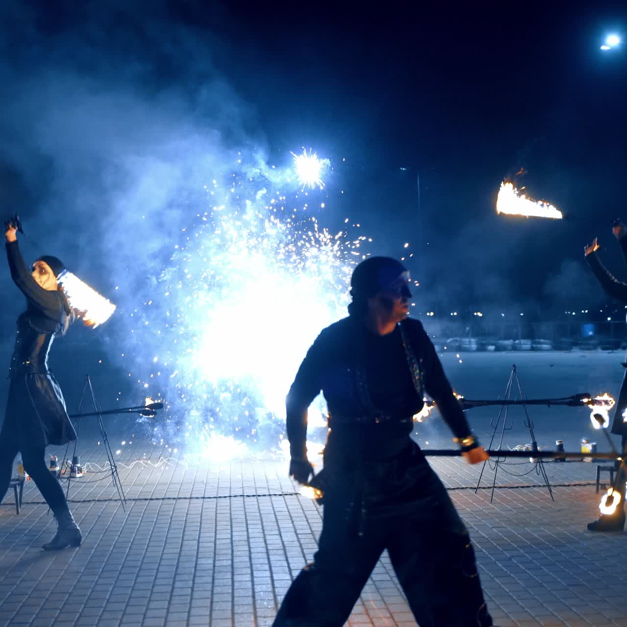 Un hombre con un disfraz negro baila haciendo girar un palo ardiendo. Dos bailarinas hacen girar los accesorios en el fuego al fondo.