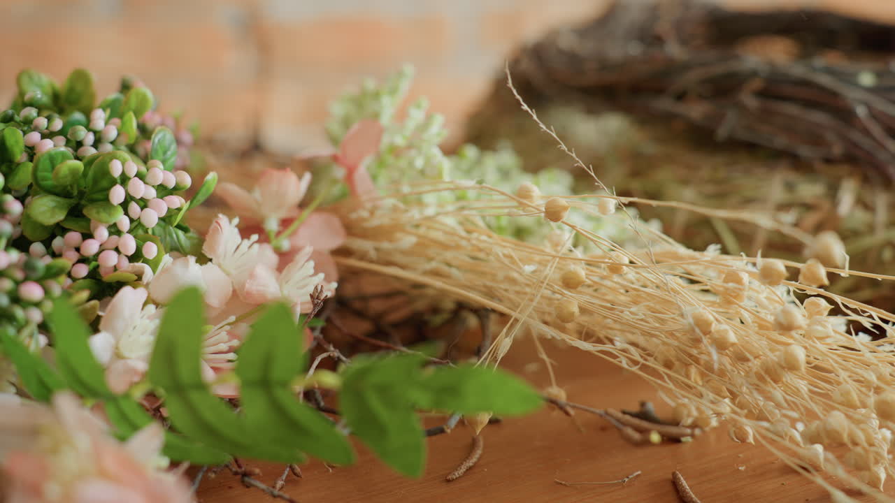 Hands arranging delicate pink blossoms, green foliage, beige dried stems, and small white flowers with straw wreath base on wooden table during creative floral decoration project