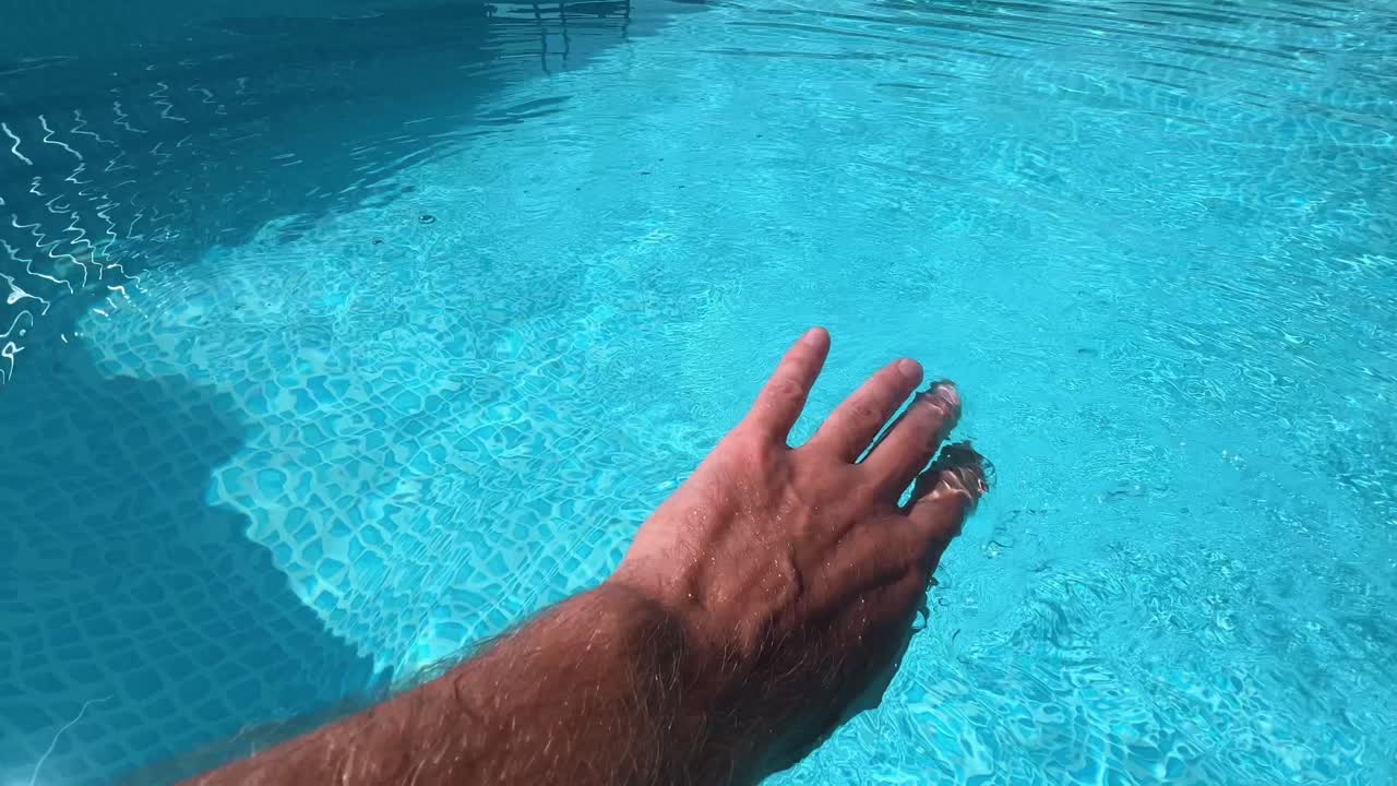 Hairy man's hand gently touching and feeling the refreshing blue water in a swimming pool, embodying the essence of summer vacation and the joy of relaxation under the sun