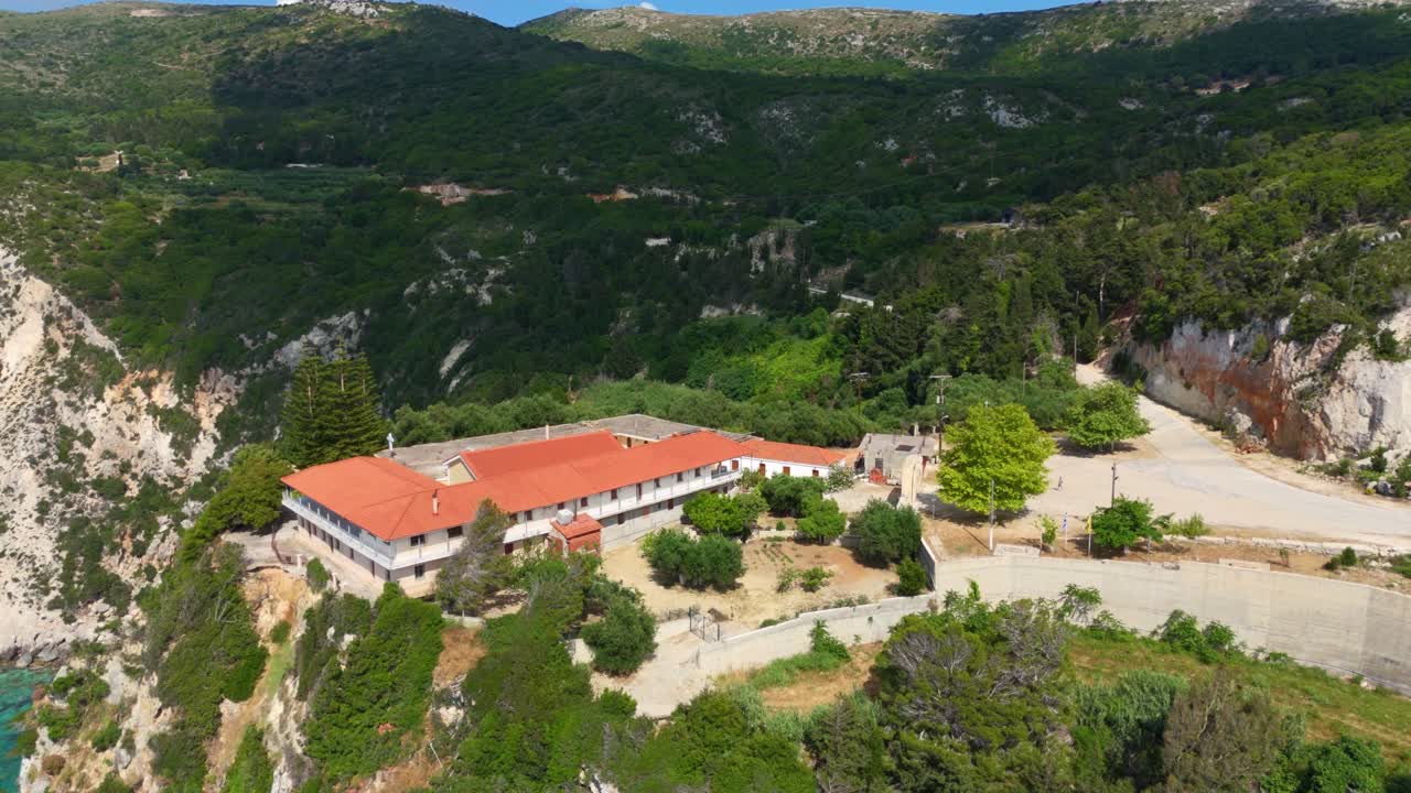 Breathtaking aerial drone view of the Monastery of Kipoureon in Kefalonia, Greece, perched on dramatic cliffs overlooking the stunning blue waters of the Ionian Sea, with rugged mountains.