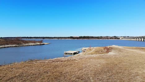 Flying-close-to-the-dormant-grass,-moving-towards-the-lake-and-a-small-inlet-which-has-a-small-fishing-dock