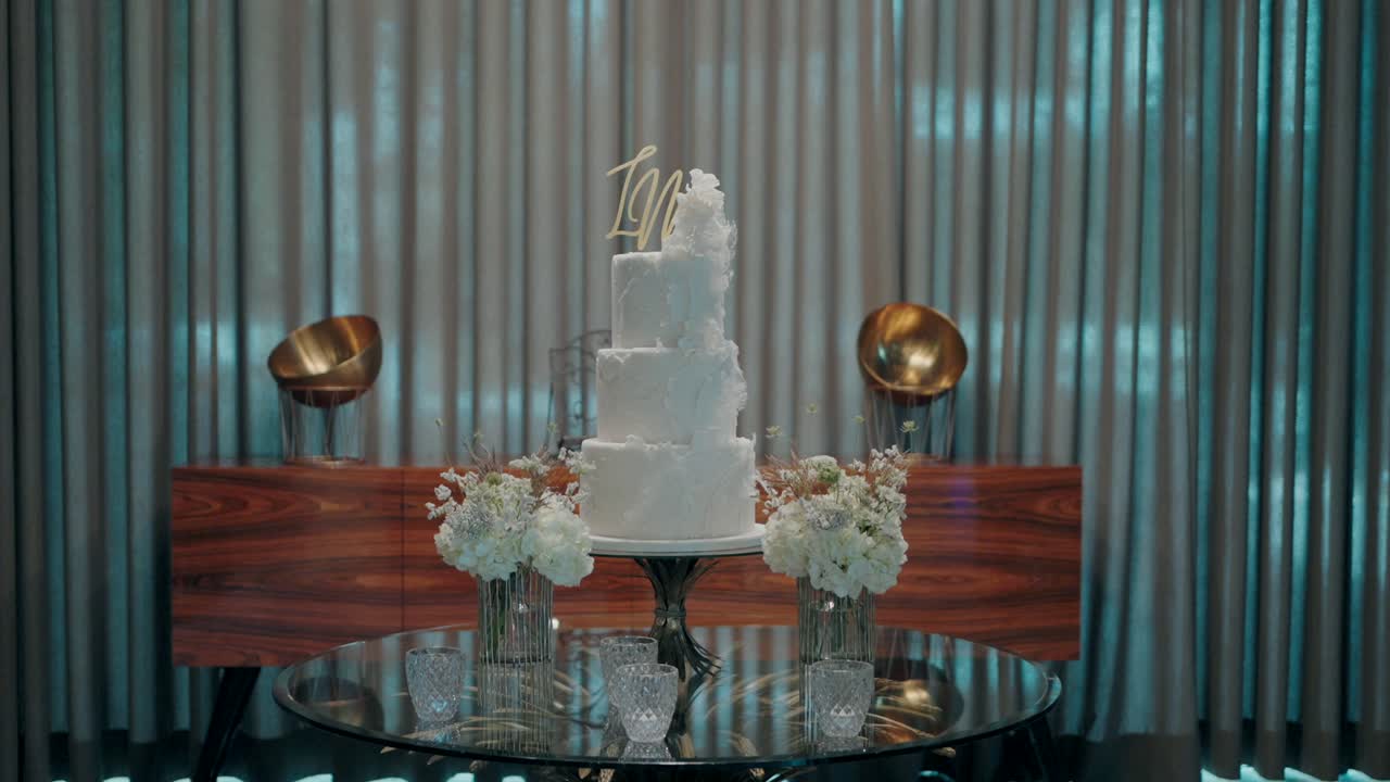 pastel de boda blanco de cuatro niveles con jarrones florales en una mesa de vidrio reflectante en un entorno sofisticado