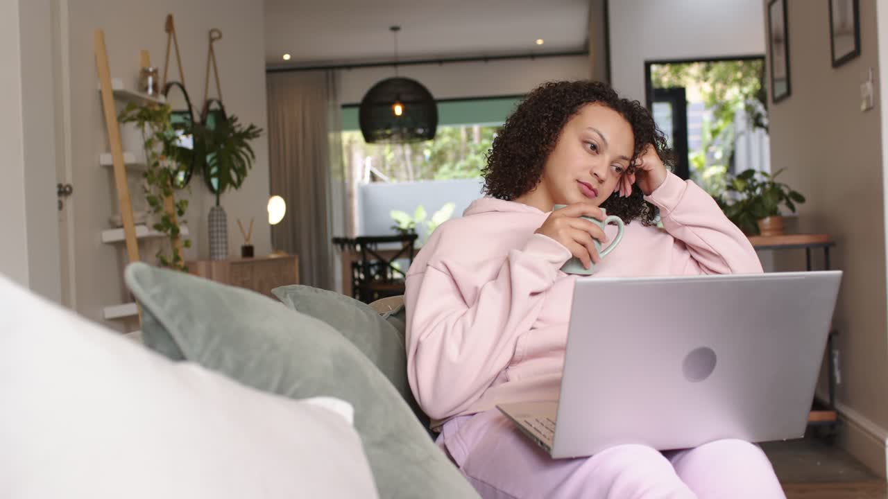 African American woman reclining on couch in pink sweatshirt, studying on laptop while sipping mug