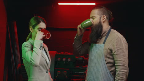 mujer de negocios caucásica parada en un taller metalúrgico con un trabajador de la industria metalúrgica con barba y delantal hablando y bebiendo té o café