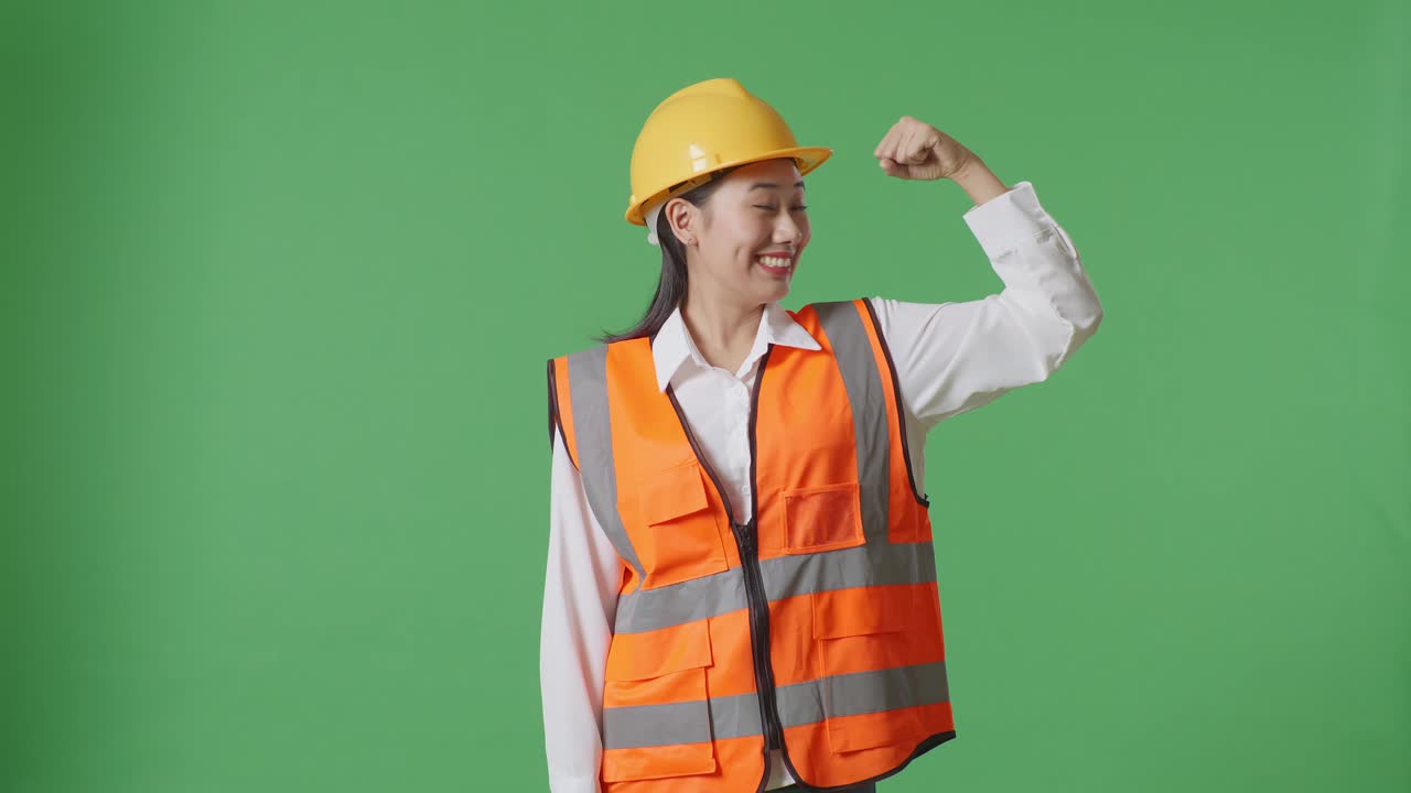 안전 헬을 입은 아시아 여성 엔지니어가 초록색 스크린 배경 스튜디오에 서서 카메라를 향해 미소 짓고 있습니다.