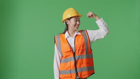ingeniera asiática con casco de seguridad flexionando su bíceps y sonriendo a la cámara mientras está de pie en el estudio de fondo de la pantalla verde