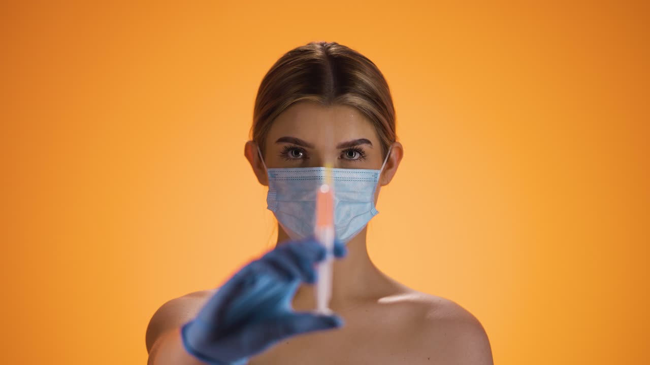 toma estática de una joven mujer bonita o enfermera mirando a la cámara y sosteniendo una jeringa con medicina mientras lleva una máscara ffp2 contra la corona y otras enfermedades frente a un fondo naranja
