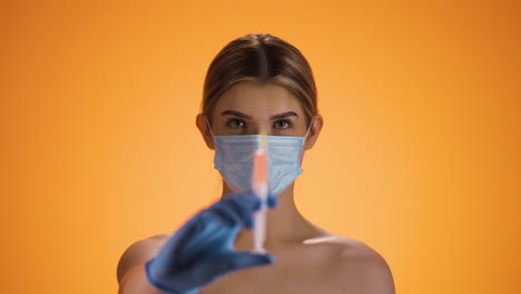 toma estática de una joven mujer bonita o enfermera mirando a la cámara y sosteniendo una jeringa con medicina mientras lleva una máscara ffp2 contra la corona y otras enfermedades frente a un fondo naranja
