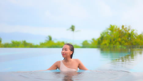 Fit-sexy-woman-relaxing-inside-the-outdoor-swimming-pool,-exotic-spa-hotel-wearing-one-shoulder-swimming-suit-on-lush-greenery-background-in-Bali