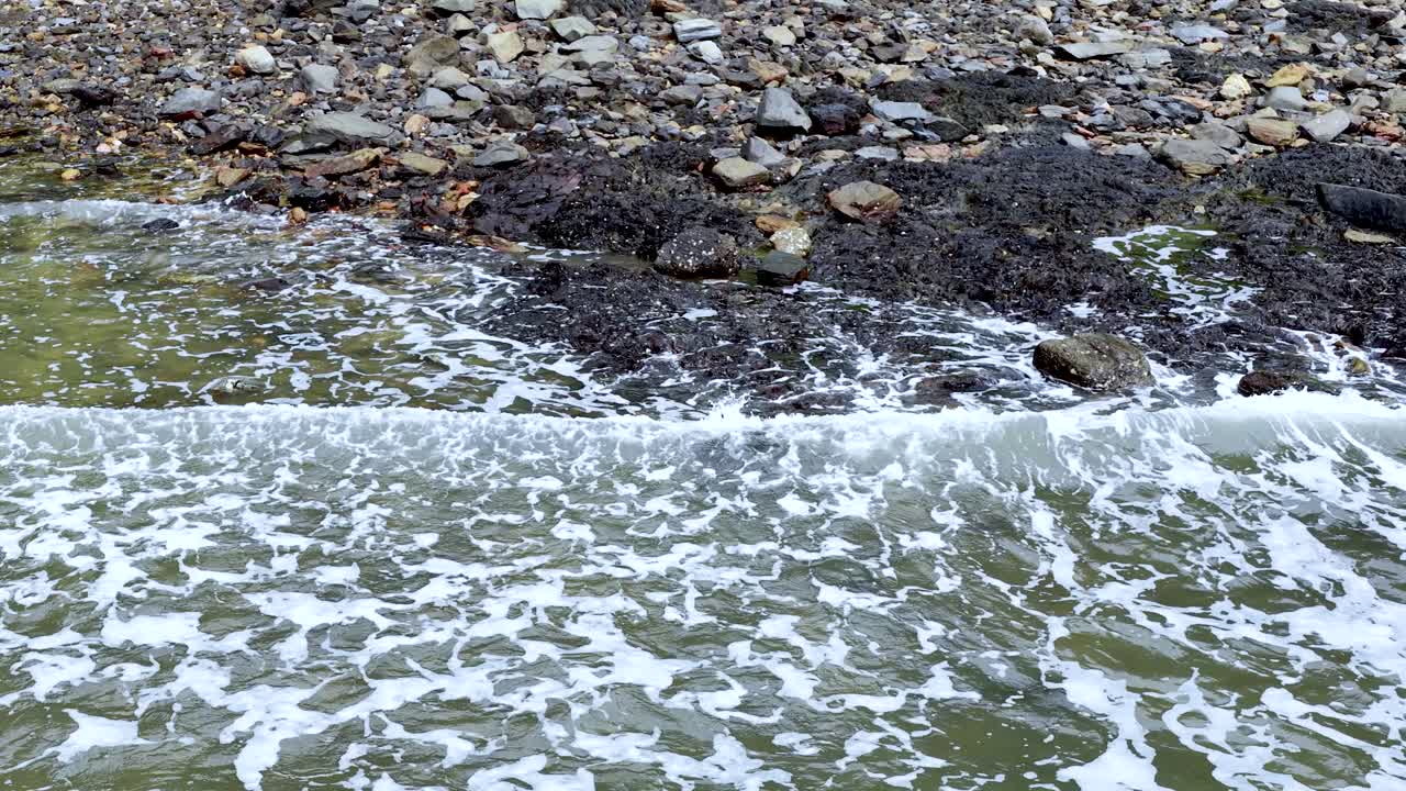 Gentle ocean waves wash over rocky shoreline, foaming water receding, natural daylight, static overhead view