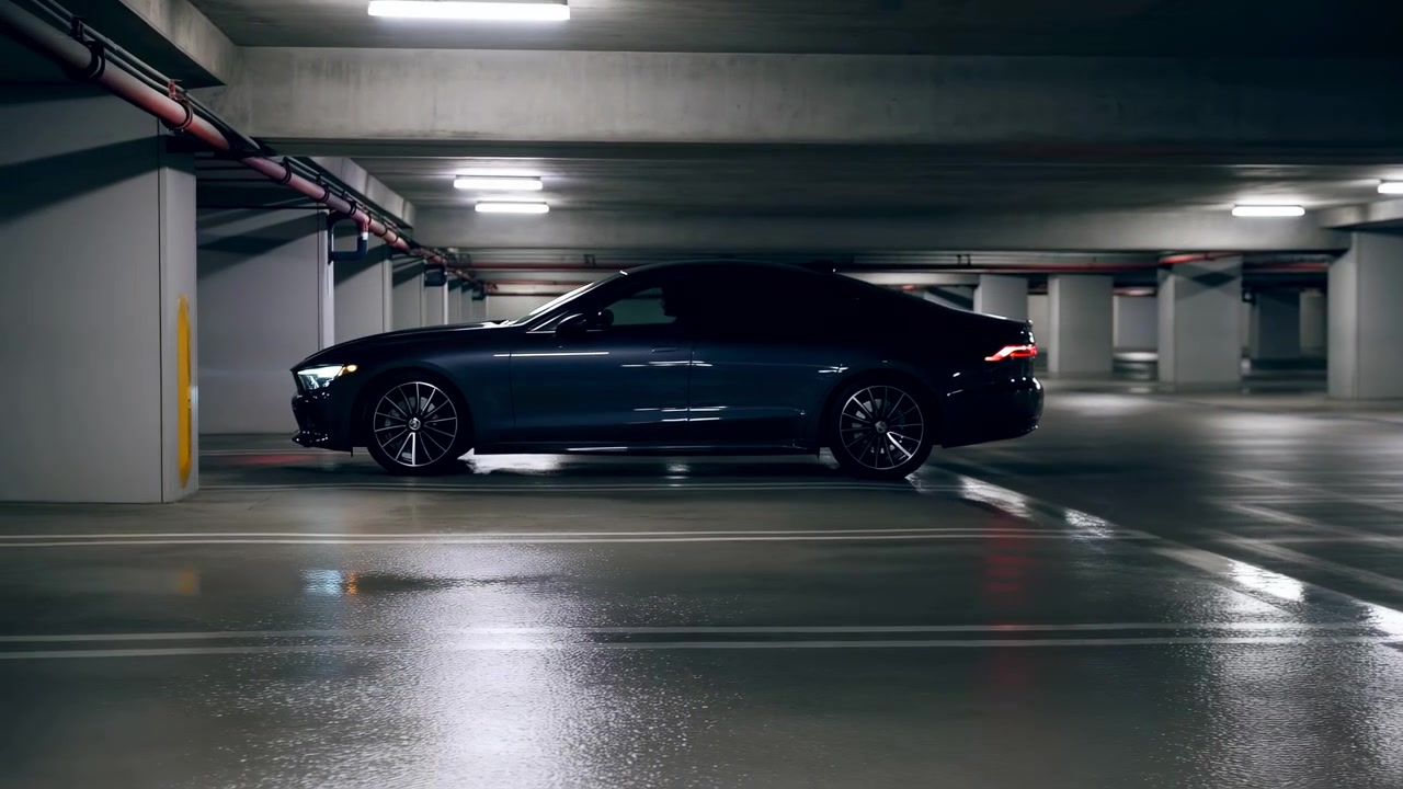 Berlina nera parcheggiata in un garage sotterraneo, vista da un'angolazione posteriore laterale con fanali posteriori luminosi e riflessi sul pavimento di cemento sotto illuminazione fluorescente.
