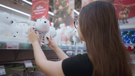 side view of young woman with long brown hair wearing black shirt and glasses admiring white plush polar bear toy with scarf on store shelf surrounded by festive holiday decorations