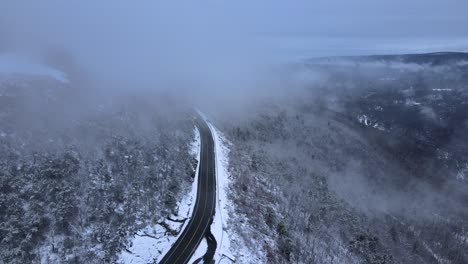 flying through clouds over snow-covered mountains and forests in a mountain valley with a scenic highway below, on a cloud winter’s day