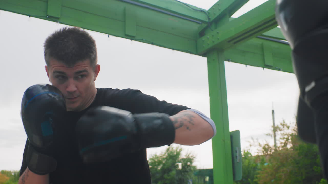 Joven boxeador caucásico practicando sparring defensivo bajo un pabellón verde con guantes de entrenamiento y guantes de boxeo, mirada concentrada, cielo nublado, bloqueos a cámara lenta y ejercicios de contraataque en un parque al aire libre.