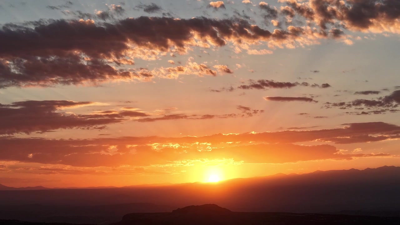 Timelapse 4K UHD con drone che cattura un'intensa alba arancione. Il sole brillante sorge su un paesaggio montano del sud-ovest in silhouette, illuminando nuvole mattutine sparse in un cielo vibrante e luminoso