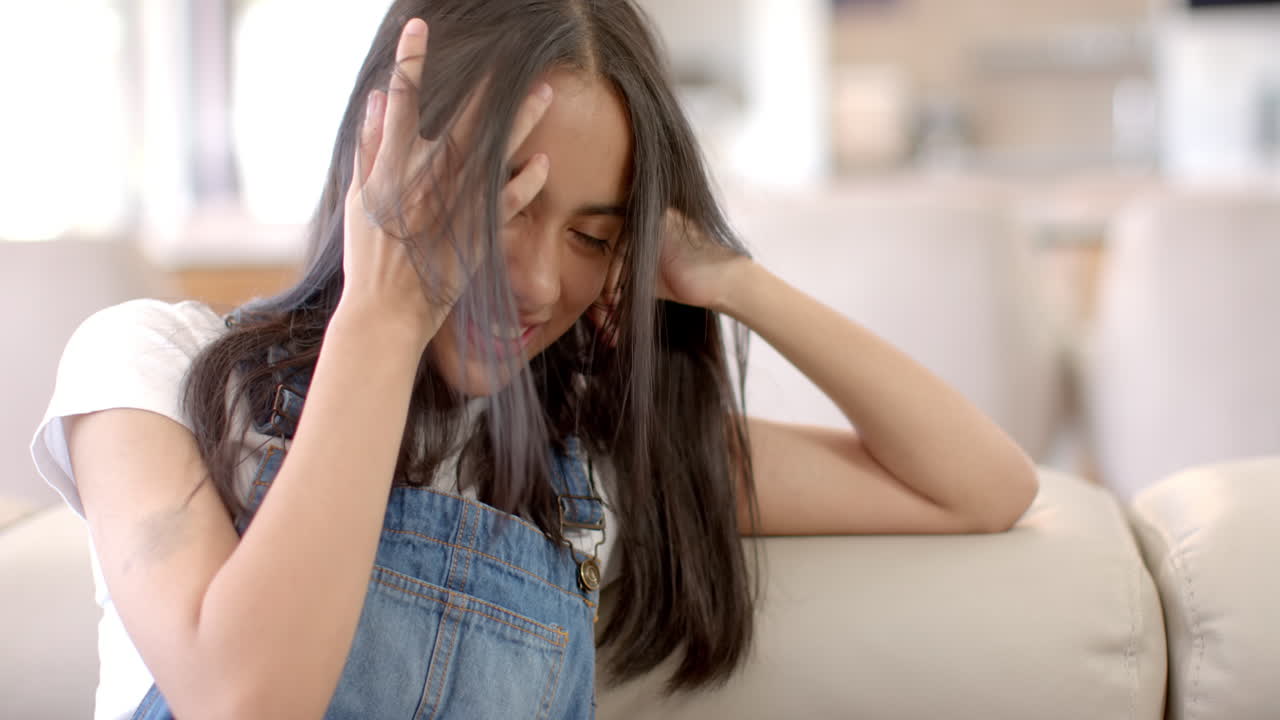 Biracial teenage girl sitting, touching hair, smiling at home