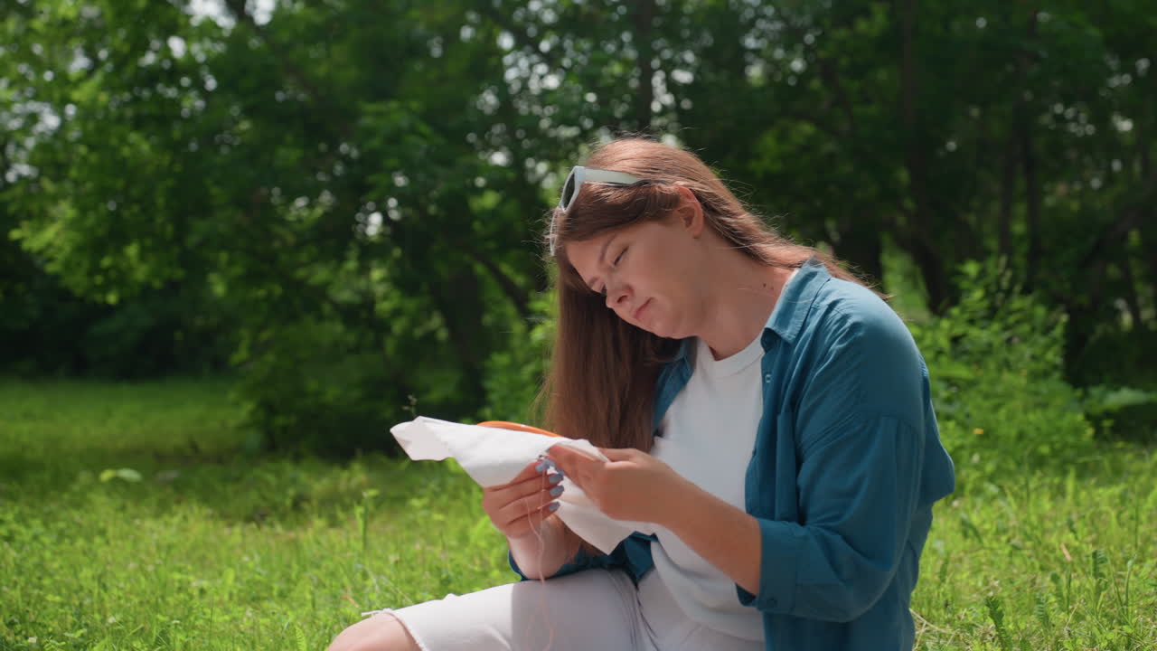 Vista laterale di una giovane donna che ricama alla luce del sole, seduta su una coperta in un tranquillo parco verde, concentrata sul delicato lavoro di cucito, circondata da alberi rigogliosi e dalla natura calma