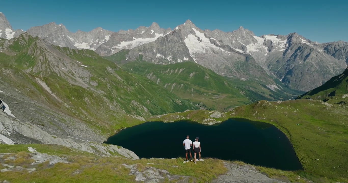 Drone approaching an athletic hiking couple standing above an alpine lake, revealing breathtaking glaciers, towering mountain peaks and a crystal-clear blue summer sky in Switzerland.