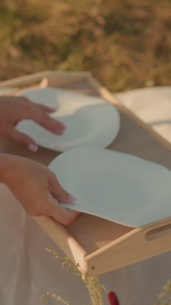 mujer sentada en una alfombra al aire libre con flores en una canasta tejida cerca, arreglando platos en la mesa de picnic bajo la luz del sol, las manos ajustando platos, creando una atmósfera de picnic pacífica e idílica en un día cálido