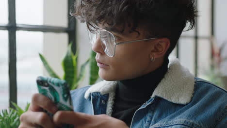 Un-Joven-Hispano-Elegante-Usando-Un-Teléfono-Inteligente-En-Casa,-Un-Estudiante-Navegando-Por-Mensajes-De-Redes-Sociales-Enviando-Mensajes-De-Texto-Por-Teléfono-Móvil-Disfrutando-Compartiendo-Un-Estilo-De-Vida-Moderno-En-Línea-Usando-Gafas