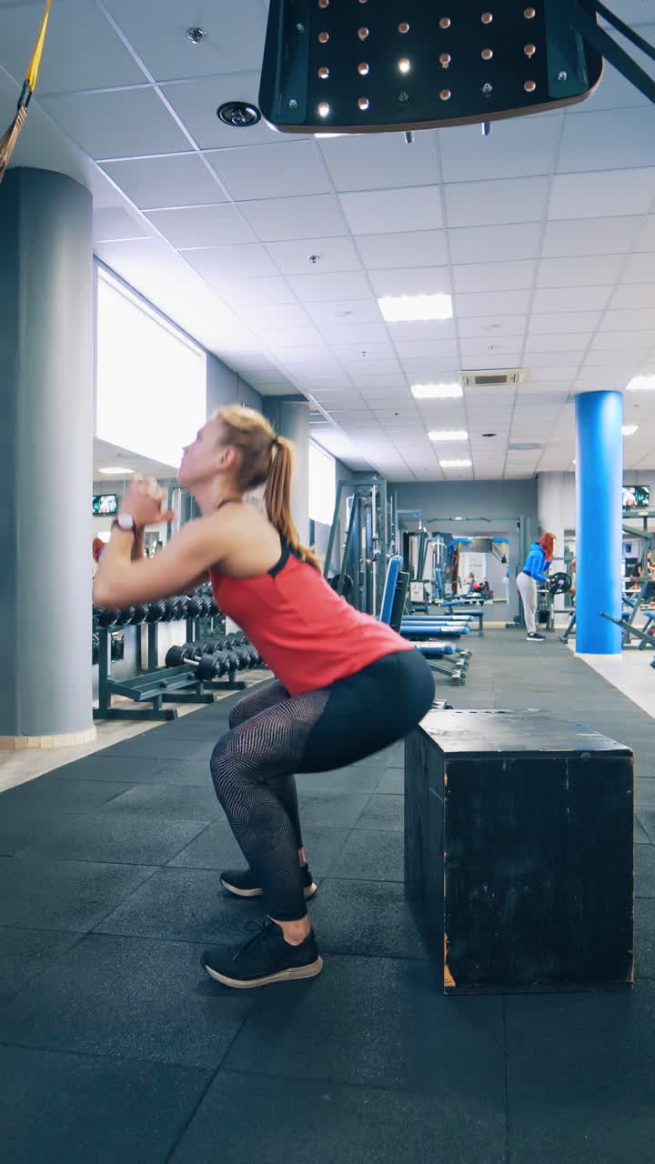 Side view of a girl in a sports club. Young woman doing squats near the wooden box in the modern fitness centre. Healthy lifestyle. Vertical video