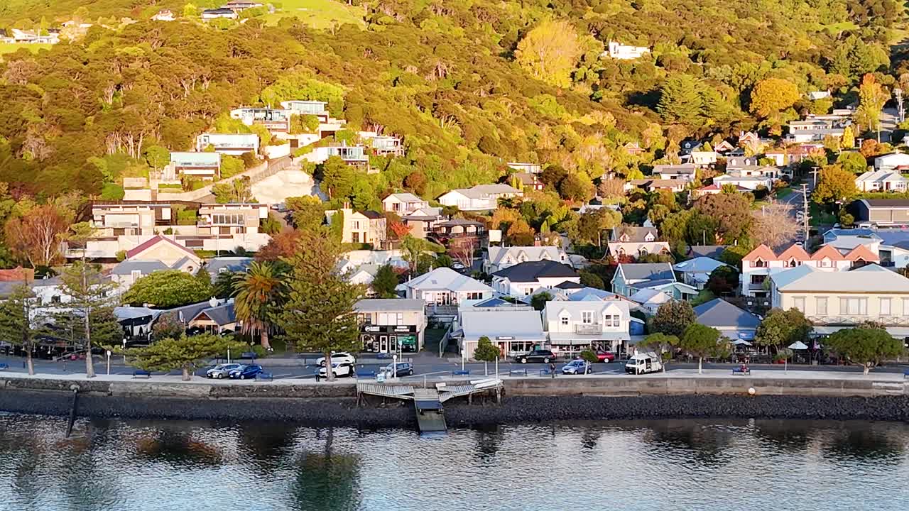 Drone footage captures Akaroa's picturesque houses and waterfront during golden hour, highlighting vibrant colors and serene atmosphere